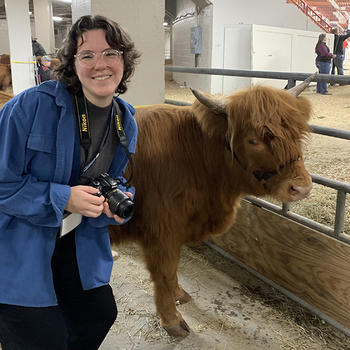 A young woman with dark hair wearing a dark shirt and a camera around her neck smiles as she poses next to a cow at the Pennsylvania Farm Show.